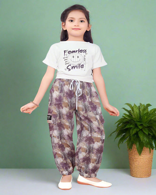 A young girl wearing a stylish co-ord set featuring a white "Fearless Smile" graphic t-shirt and magenta-brown printed jogger pants with a drawstring waist. She is posing confidently indoors with a modern, elegant background. Perfect casual wear for kids.